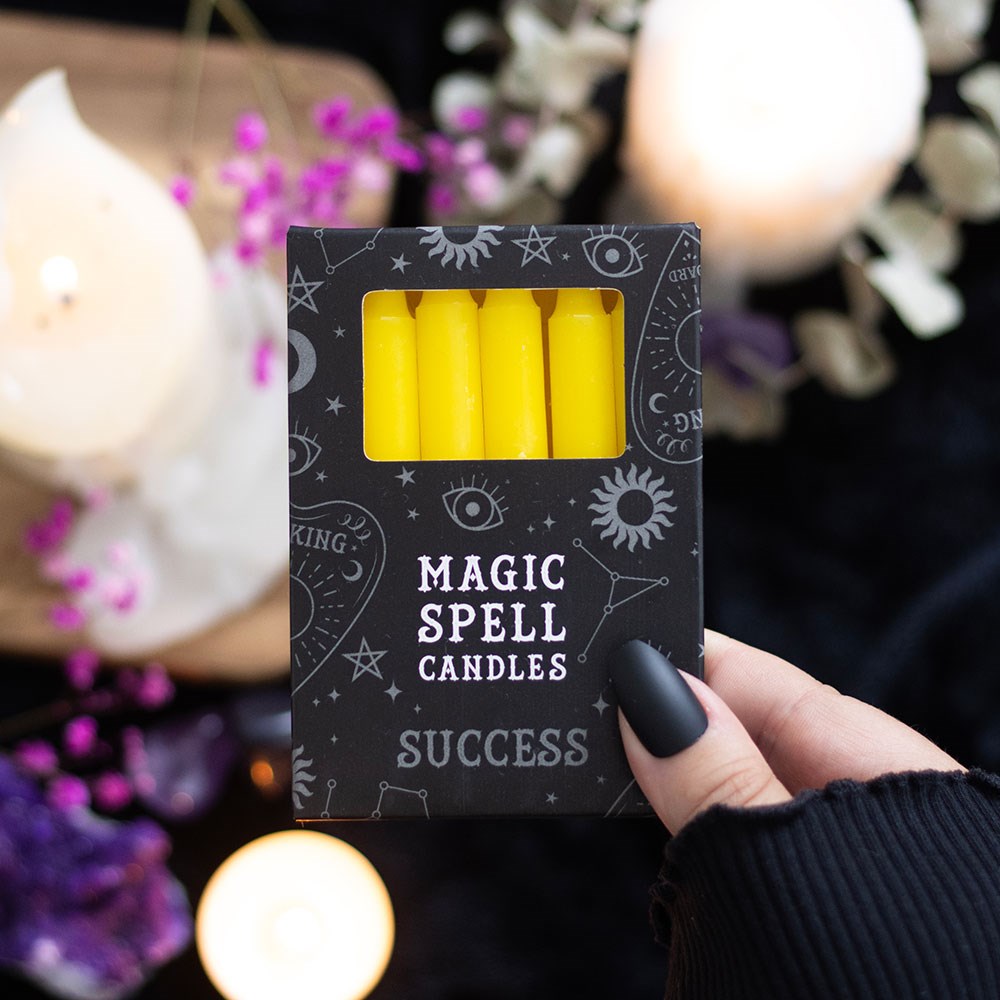 Box of 'Magic Spell Candles' labeled 'Success' held by a hand with a blurred candle and flowers in the background.